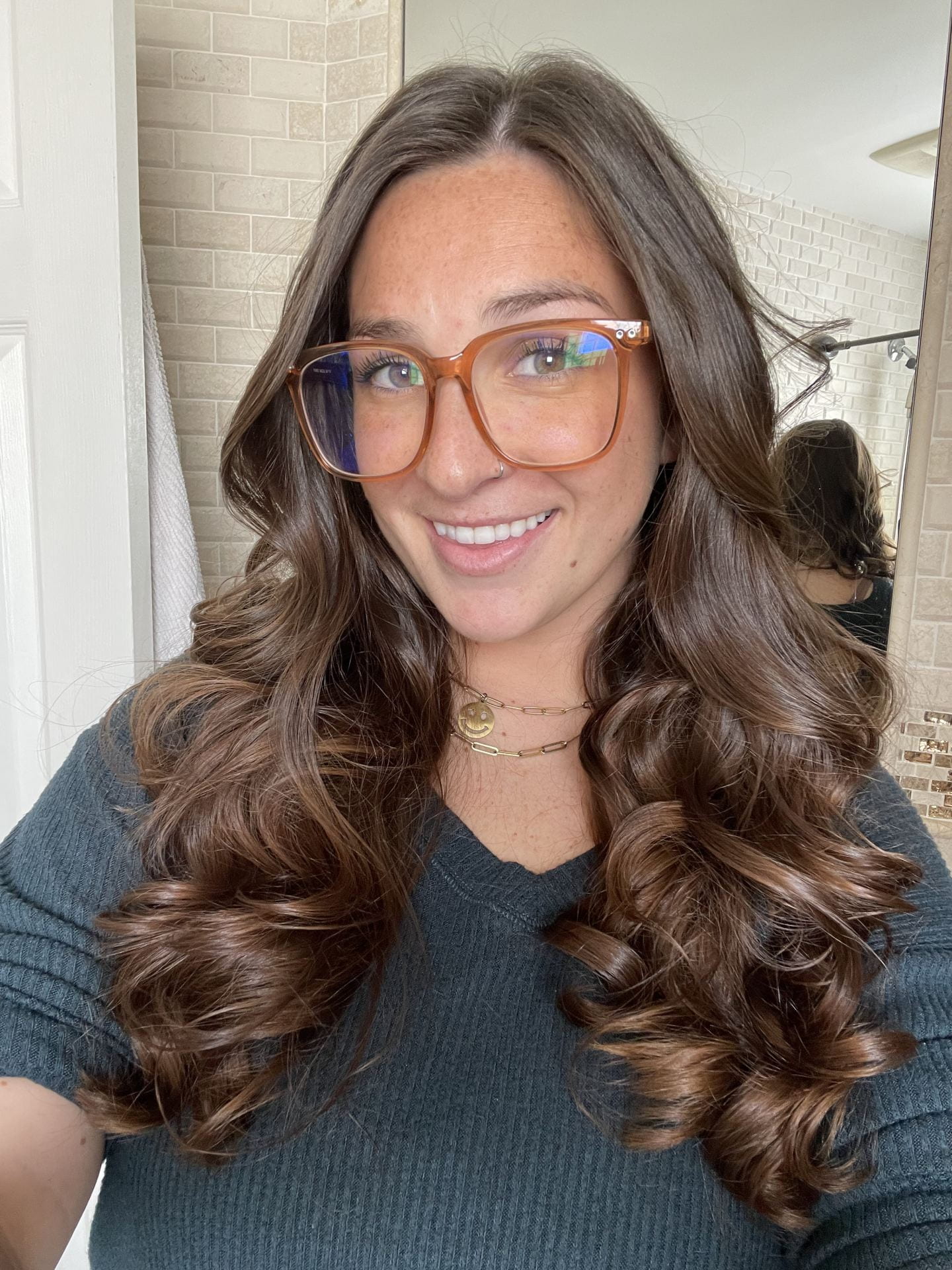 A smiling young person with long brown hair, glasses, and a necklace, wearing a blue-green shirt.